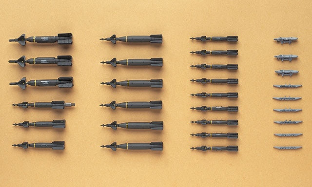 A set of miniature model aircraft or missiles arranged in rows on a warm tan background. The items are primarily dark gray with black and gold accents, alongside several smaller light gray pieces on the right.